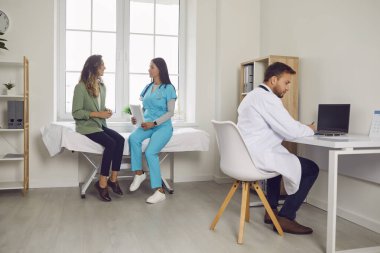 Gülümseyen kadın hasta, klinikteki tıbbi muayene sırasında kanepede oturan genç ve dost canlısı bayan hemşireyle konuşurken, erkek doktor da ofisteki masada oturmuş tedavi öneriyor..