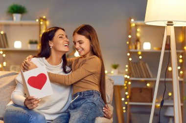 Mutlu kadın ve sevgi dolu çocuk Anneler Günü 'nde arka planda rahat bir şekilde kanepede yan yana oturup eğleniyorlar. Kağıt kartla mutlu bir anne kızını kollarında tutuyor. 