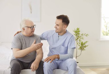 Klinikte muayene ettikten sonra kıdemli bir adamla konuşan cana yakın genç bir hemşire. Hasta bakıcı ile hastanede yatağında oturan yaşlı hasta. Hemşirelik hizmeti ve destek kavramı.