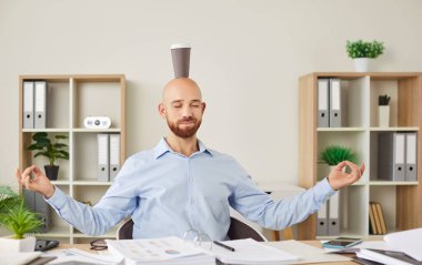 Sakin, sakin bir ofis çalışanı masasında oturmuş kafasında fincanla yoga Mudra yapıyor, stresi azaltmak için meditasyon yapıyor, rahatlama yorgunluğu çekiyor, iş yerinde dengeyi kuruyor, zihinsel sağlığını geliştiriyor.