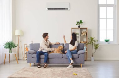 Genç bir çift, kanepede köpekli mutlu bir aile, klimayı açıyorlar, uygun ev ortamı için sıcak, soğuk hava sıcaklığı kontrolü sağlıyorlar, ideal iklim kontrol sistemi sağlıyorlar.