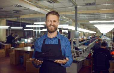 Portrait of proud shoe factory worker with new product. Happy handsome bearded man standing in workshop, holding leather boot, looking at camera and smiling. Footwear manufacturing industry concept clipart