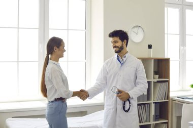 Sağlık ve ilaç servisi. Dost canlısı gülümseyen genç erkek doktor, muayenehane toplantısında kadın hastaya selam veriyor ve el sıkışıyor. Ortaklık, güven ve tıbbi etik kavramı.