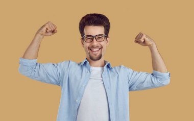 Sıradan beyaz tişörtlü, bej arka planda mavi gözlüklü, gülümseyen mutlu sakallı adamın portresi kaslarını gösteriyor ve kendini güçlü hissediyor. Spor, liderlik, güç kavramı.