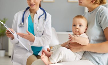 Kliniği ziyaret ederken kucağında yeni doğmuş bir bebeği tutan genç bir anne resmi. Dost canlısı bayan çocuk doktoru, çocuğun sağlık muayenesinden sonra bebeğe bakan anneye danışmanlık yapıyor..