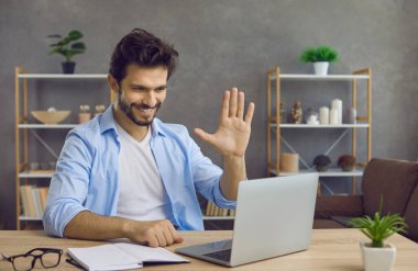 Masasında dizüstü bilgisayarla oturup gülümseyen ve ekranda el sallayan yakışıklı genç adam. Üniversite öğrencisi ya da uzaktan çalışan bir şirket çalışanı video çağrısına başlarken öğretmenini ya da meslektaşını selamlıyor.