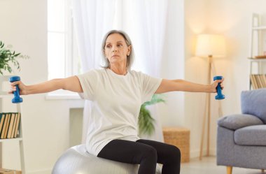 Spor kıyafetleri giyen yaşlı bir kadın evde spor egzersizleri yapıyor. Oturma odasında halterler üzerinde oturuyor. Emekli kadın içeride egzersiz yapıyor. Spor, spor ve spor konsepti.