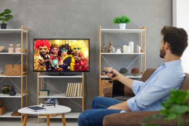 Adam film izliyor ya da evde TV şovu yapıyor. Genç adam kanepede oturuyor ve televizyonu değiştiriyor. Modern oturma odasının iç dekorasyonunda duvarların üzerinde duran büyük HD ekranla.