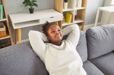 Genç Afrikalı Amerikalı kadın ellerini başının üstüne koyup rahat gri koltukta dinleniyor. Gülümseyen siyah kız ara veriyor, işten dinleniyor ya da ders çalışıyor, evde boş vakit geçiriyor..