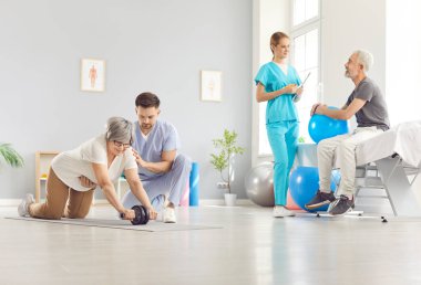 Hemşire ve fizyoterapist rehabilitasyon kliniğinde yaşlı erkek ve kadına yardım ediyor. Fizik tedavi gören yaşlı çiftler, halterlerle spor egzersizleri yapıyorlar ve sağlık ve sağlık için yoga topu kullanıyorlar..