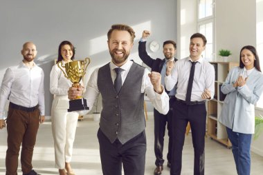 Ofiste iş adamlarıyla çevrili mutlu bir işadamı, bir şampiyonluk kupası tutuyor, bir başarıyı veya başarıyı kutluyor. Meslektaşlar alkışlıyor, tebrik ediyor, destekliyor ve birlikte..