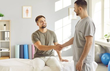 Erkek kayropraktik uzmanı veya fizyoterapist başarılı bir tedaviden sonra rehabilitasyon hastanesinde kanepede oturan bir adamla el sıkışıyor. Fizyoterapi ve osteopatik tıp konsepti.