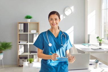 Ofisinde mavi üniformalı gülümseyen bir kadın doktorun portresi. Kendine güveni tam bir doktor. Raporu randevusuyla birlikte tutuyor ve kameraya bakıyor..