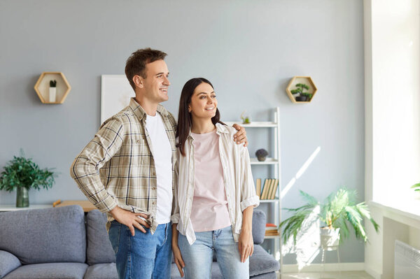 Портрет молодой счастливой улыбающейся пары, стоящей в гостиной и смотрящей в окно вместе. Улыбающиеся семейные мужчина и женщина обнимаются в своем доме. Понятие любви и гармоничных отношений.