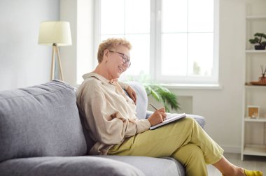 Mutlu gülümseyen olgun kadın portresi. Günlük giysiler içinde. Oturma odasındaki kanepede oturuyor ve not defterine notlar yazıyor. Kendinden emin son sınıf öğrencisi, liste planlaması için yazıyor..
