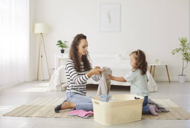 Anne ve küçük çocuk birlikte çamaşır yıkıyorlar. Kadın ev hanımı ve kız çocuğu çamaşır sepetiyle yerde oturuyor, temiz giysileri katlıyor, ya da çamaşır yıkamadan önce kirli çamaşır ve kumaşları renklerine göre sıralıyor.