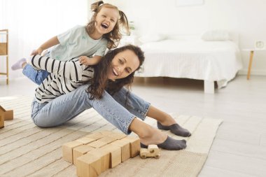Oturma odasında, yerde oturup ahşap oyuncaklarla oynayan tatlı bir kızla eğlenen mutlu genç annenin portresi. Gülümseyen kadın ebeveyn kızıyla vakit geçiriyor..