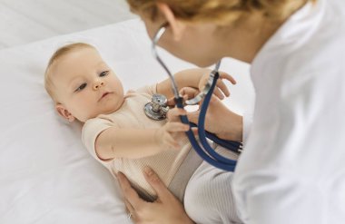 Stetoskop kullanan, sağlık muayenesinde bebeğinin kalp atışlarını dinleyen bir kadın doktor görüntüsü. Kanepede yatan bebeği muayene eden profesyonel bir çocuk doktoru..