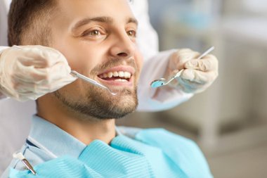 Modern dişçilik kliniğinde tıbbi muayene sırasında ağzı açık bir erkek hastanın yakın plan portresi. Dişçi, dişçi aletleriyle genç adamın dişlerini inceliyor. Diş sağlığı hizmetleri.