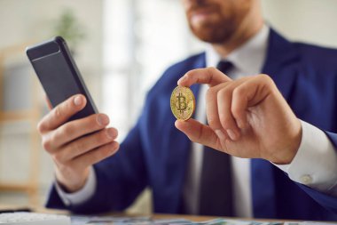 Ofisteki masada elinde cep telefonu ve bitcoin ile oturan başarılı borsa simsarı ya da iş adamı yakın plan. Para yatırımı, kripto para birimi ve yatırım konsepti.