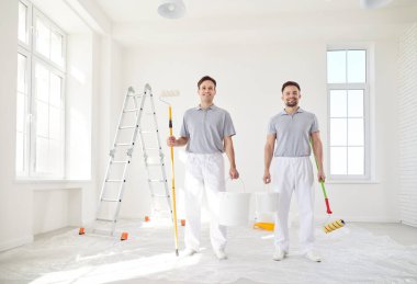 Gülümseyen erkek ressamlar ya da inşaat işçileri, profesyonel servis ekibi merdiven kenarında koruyucu üniforma giyip hazırlanmaya hazır, boya fırçasıyla resim yapıyorlar.