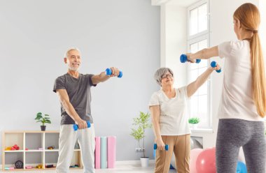 Spor malzemeleri ve eğitmenle spor yapan iki mutlu son sınıf öğrencisi. Gülümseyen yaşlı adam ve kadın spor salonunda durup antrenörlere bakıyor ve halterlerle egzersiz yapıyorlar.