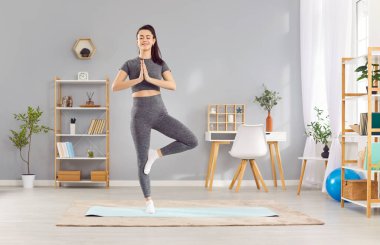 Spor kıyafetleri giyen, yoga egzersizi yapan, oturma odasında duran sakin, genç bir kadın. Gözleri kapalı beyaz bir kız evde meditasyon yapıyor. Sağlıklı yaşam tarzı ve ev eğitimi kavramı.