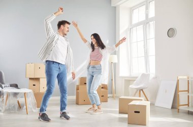 Genç çift taşınma gününü kutluyor, gülümsüyor ve karton kutularla dolu yeni bir dairede dans ediyor. Aile bir mülk almaktan mutluluk duyuyor. Taşınma, taşınma, emlak, ipotek konsepti.