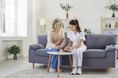 Genç, gülümseyen hemşire kadın, evdeki kanepede oturan tahta yapı taşlarıyla gri saçlı yaşlı bir adamla oynuyor. Bakıcı desteği, sağlık hizmetleri ve emeklilik konseptinde boş zaman.