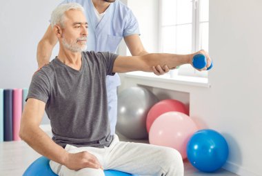 Doktorun ya da fizyoterapistin, yaşlı erkek hastaya dambılla egzersiz yaptırmasına yardım etmesi. Klinikte kol rehabilitasyonu sırasında yetişkin bir adamı destekleyen erkek bakıcı.