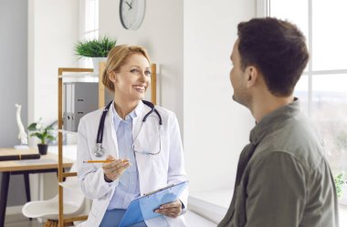 Tıbbi ofiste oturan hasta bir gencin portresi randevu ile rapor tutan ve klinikteki tıbbi muayene sırasında danışmanlık yapan dost canlısı bir kadın doktoru dinliyor..