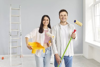 Genç çift mutlu ressamlar yeni bir daire inşaatı planlıyorlar, boya fırçasıyla duvar boyuyorlar, koruyucu üniforma giyiyorlar oda taşıma, bina onarımı, yenileme ve yenileme işleri için görev yapıyorlar.