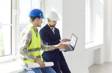 Mimar, mühendis veya iç mimar ustabaşı tasarım projesini veya yeni dairenin tamirini pencerenin yanında dikilirken, başlık takarak ve yenileme planını tartışırken dizüstü bilgisayar ekranında gösteriyor.