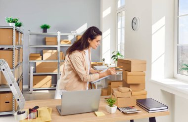 Genç bir kadın, küçük bir depodaki iş yerindeki karton kutuları inceliyor. Kadın küçük işletme sahibi envanter yapıyor, siparişleri kontrol ediyor ve panoya not alıyor. Çevrimiçi dükkanda stok envanteri.