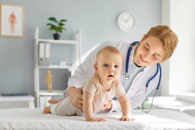 Gülümseyen genç aile doktoru, iki eli klinikte kanepede yatan küçük bebeği destekliyor. Neşeli çocuk doktoru, çocuk sağlığı kontrolü sırasında bakım ve koruma sağlıyor. Annelik kavramı.