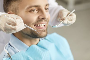 Profesyonel diş aletleriyle dişçi doktoru tarafından muayene edilen gülümseyen genç adamın yakın plan çekimleri. Erkek hasta diş hekimliği kliniği ziyareti sırasında düzgün bir ağız bakımı sağlıyor. Diş bakımı ve tedavisi