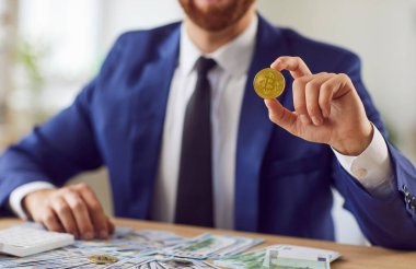 Masada oturan takım elbiseli bir erkek borsa simsarı. Elinde para, ofisinde nakit para ve elinde bitcoin var. Kripto para yatırımı konusunda başarılı bir iş adamı. Yatırım ve iş konsepti.