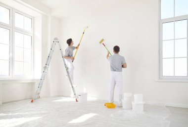 Üniformalı iki boyacı duvarları ve tavanları beyaz boyuyor. Daire yenileme konsepti. İnşaat müteahhitliği, boş bir odayı yenileme hizmeti