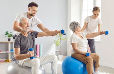 Spor malzemeleriyle fizyoterapi egzersizleri yapan mutlu yaşlılar. Gülümseyen fizyoterapistler spor topları üzerinde oturan ve dambılları tutan yaşlı adama ve kadına yardım ediyorlar.
