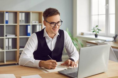 Ofiste dizüstü bilgisayarda çalışan mutlu iş adamının portresi ya da not alan ya da günlük toplantı planlayan ve bilgisayar ekranına neşeli bakan genç adam..