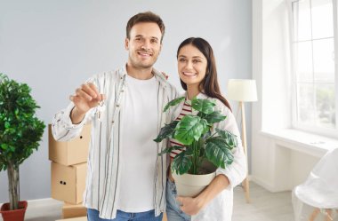 Karton kutularla çevrili aydınlık bir evde kameranın anahtarlarını gösteren neşeli genç bir çiftin portresi. Mutlu gülümseyen aile elinde çiçek saksısı ve taşınma gününde sarılıyor..