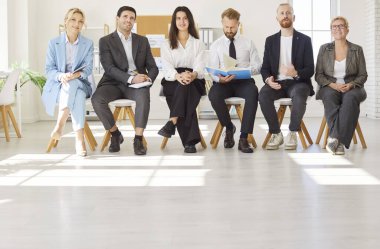 Group of smiling confident business people sitting in a row on chairs and listening to colleague or their leadership at working place during business training or conference in meeting room. clipart