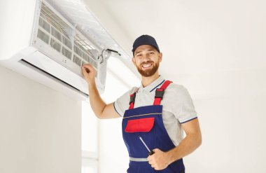 Mutlu bir teknisyenin portresi. Evdeki klimayı temizlemeye ve bakımına odaklanmış. Neşeli elektrikçi onarım ve hizmet kalitesini vurgulayarak, optimal performans ve hijyen sağlıyor.