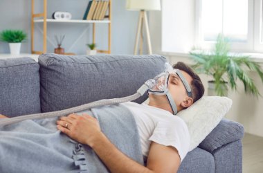 Kanepede yatan erkek hasta tıbbi yüz oksijen maskesi takıyor, ciğerler için oksijen gazı soluyor, havalandırma desteği, koronavirüs hastalığından kaynaklanan yapay akciğer havalandırması, korona virüsü zatürresi.