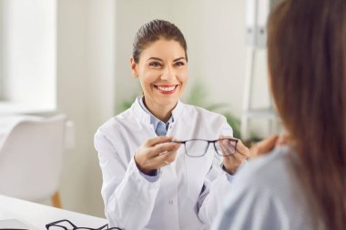 Beyaz önlük giyen profesyonel gülen gözlükçü ya da göz doktoru, görme yetisini geliştirmek için kadın hastaya yeni gözlük verir. Oftalmoloji, optometri, görüş bakımı ve göz sağlığı konsepti.