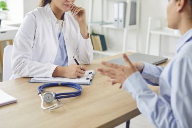 Beyaz üniformalı doktor hastane masasında oturup konuşuyor, hastaya danışıyor, pratisyen hekim klinikteki kadına danışmanlık yapıyor, sağlık yardımı, destek, randevu alıyor.