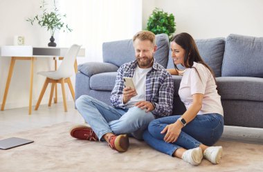 Mutlu gülümseyen bir çiftin portresi. Oturma odasındaki koltukta oturuyor ve akıllı telefon kullanıyor. Birlikte internet uygulamaları kuruyorlar. Genç adam neşeli karısına cep telefonundan video veya haber gösteriyor..