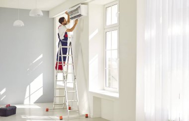 Profesyonel erkek teknisyen yeni eve modern duvar kliması kuruyor. İş üniformalı bir adam parlak, ferah bir odada merdivenin üzerinde duruyor ve klimayı tornavidayla monte ediyor..