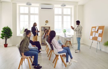 Çok uluslu iş adamlarından oluşan bir grup şirket çalışanı bir toplantıda arka arkaya koltuklarda oturuyor ve konferansta veya modern ofisteki seminerde konuşan Afrikalı Amerikalı liderleri dinliyor..