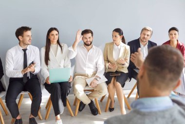 Gülümseyen genç adamın portresi bir iş toplantısında soru sormak ya da cevap vermek için elini kaldırıyor. Bir grup şirket çalışanı ya da bir grup personel konferans ya da eğitim sırasında koltuklarda oturuyorlar..
